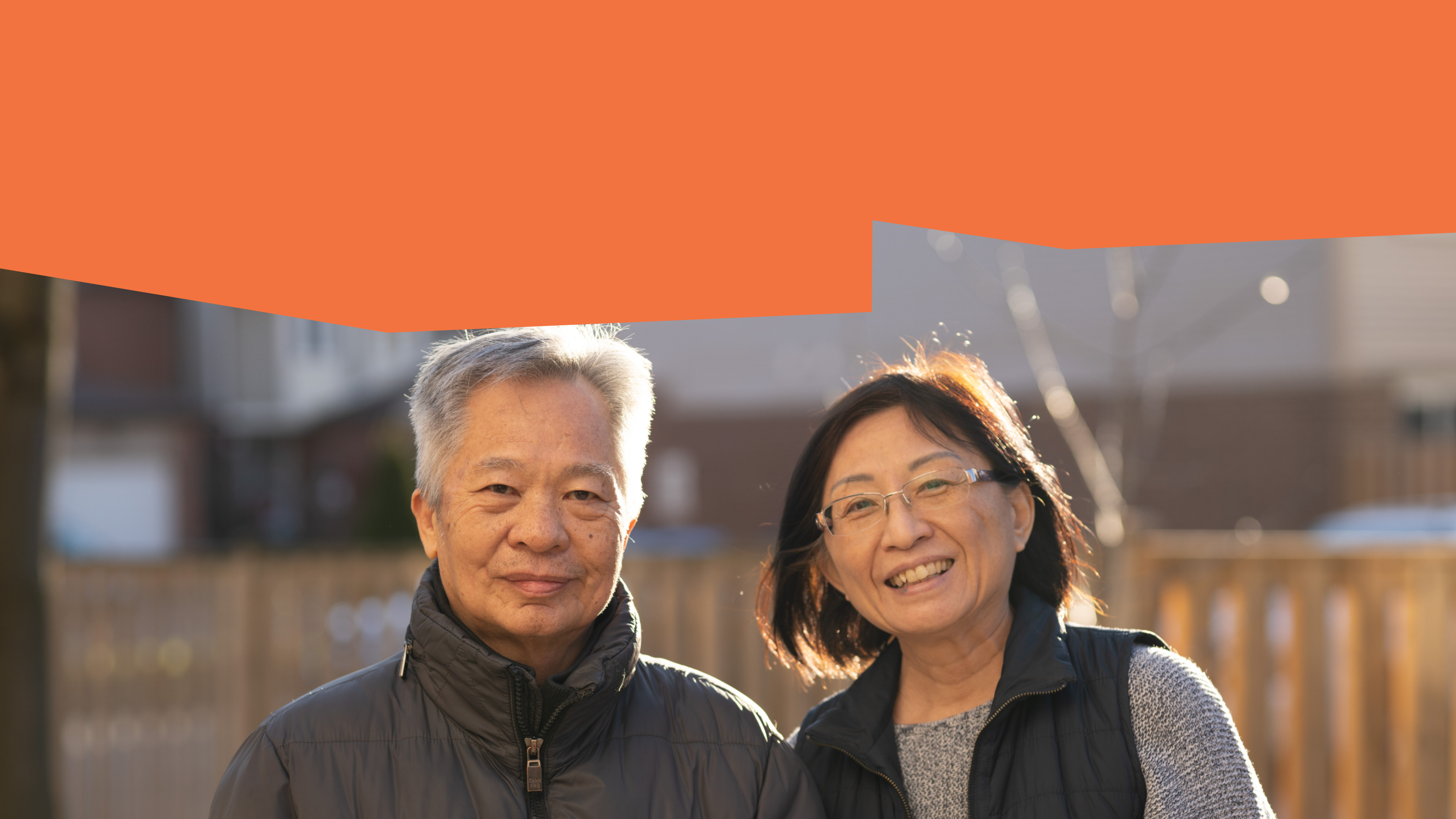 Two seniors are standing outside and smiling at the camera wearing black winter clothing. One of them has grey hair, the other has black hair and glasses.