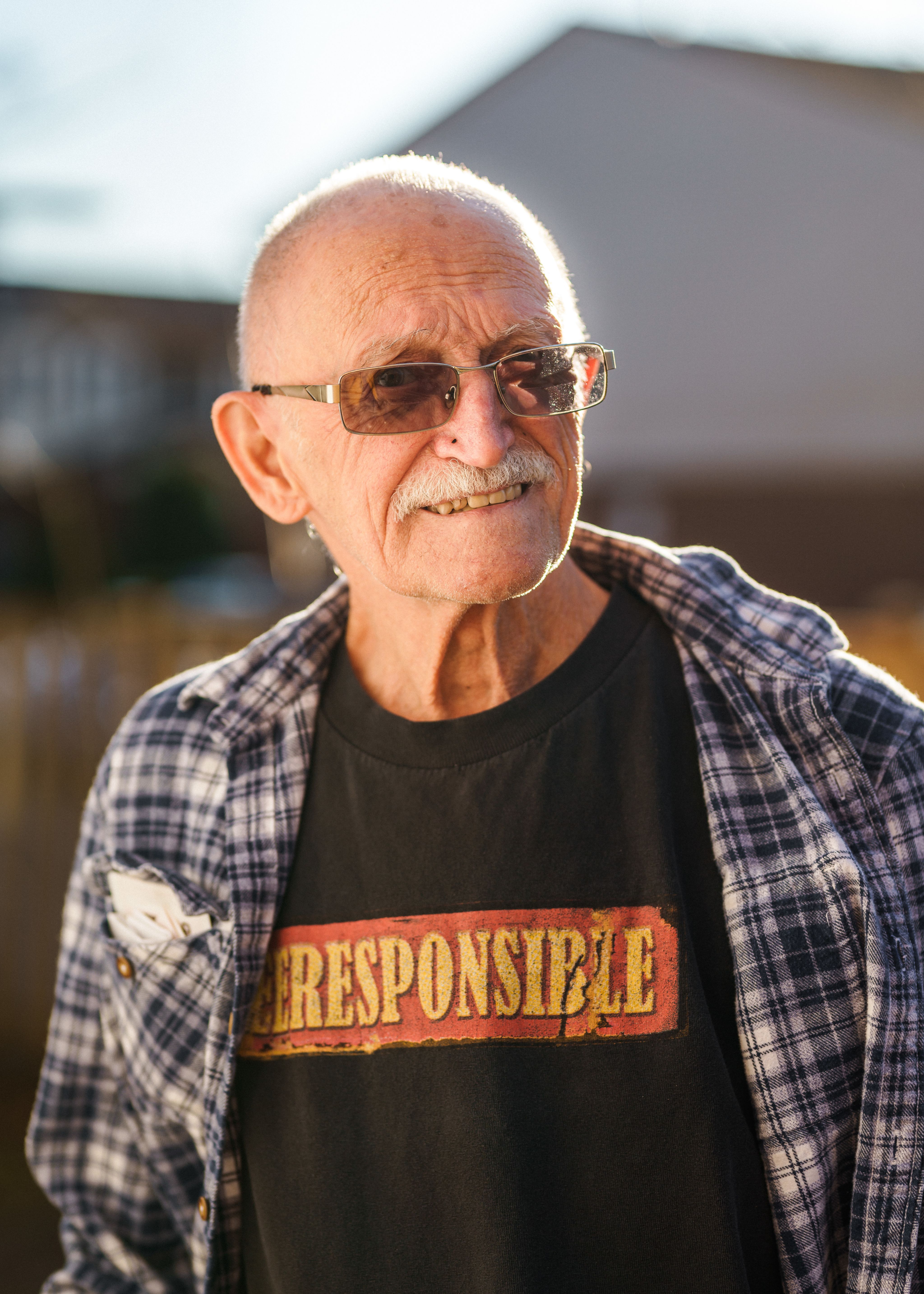 An older man with a white mustache is standing outside wearing a plaid shirt and tinted glasses.