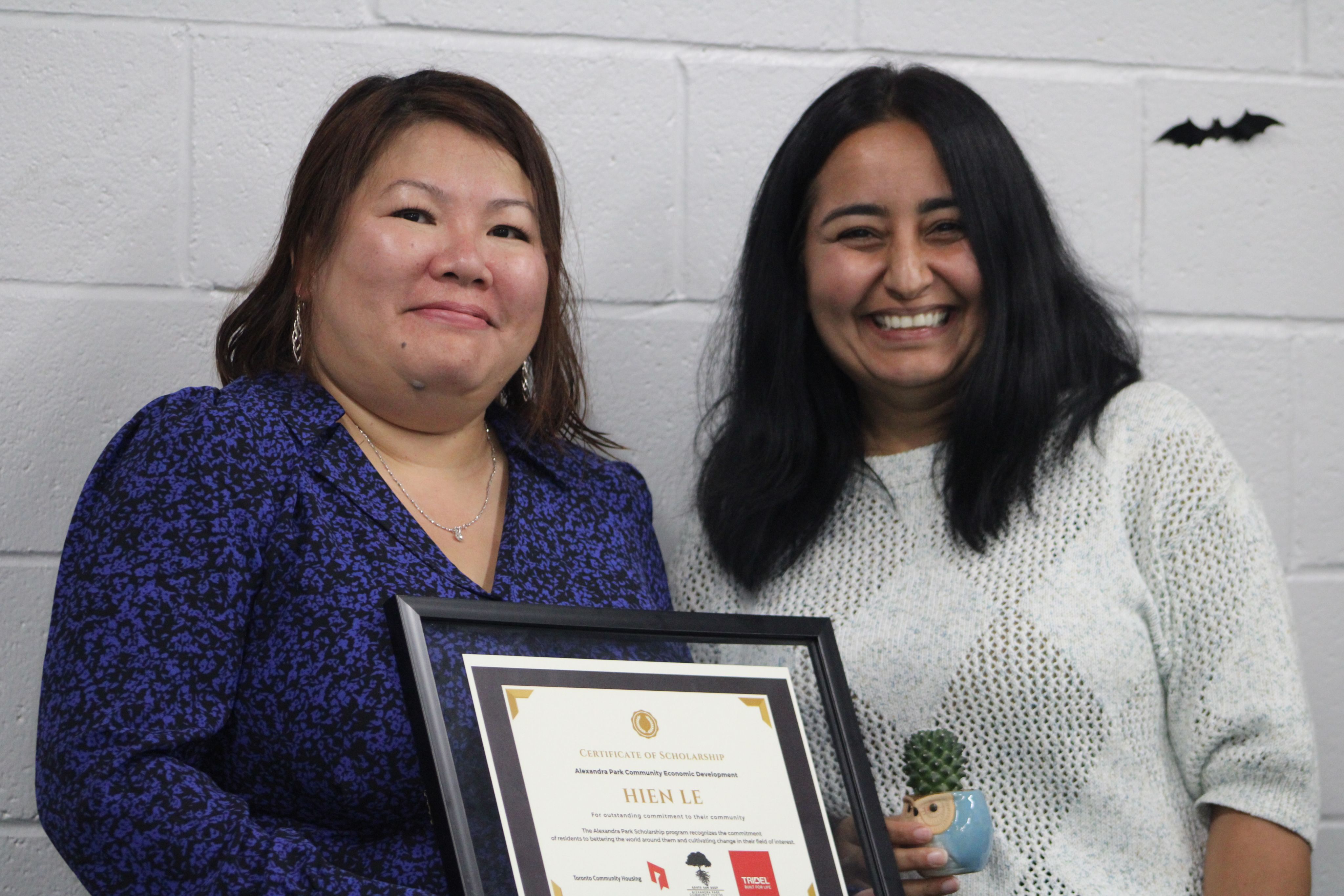 A woman wearing a blue dress is holding a framed certificate while a woman in a white sweater poses beside her, smiling.