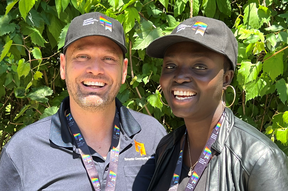 Two people posing in Pride colours wearing hats and lanyards featuring TCHC logos in Pride colours