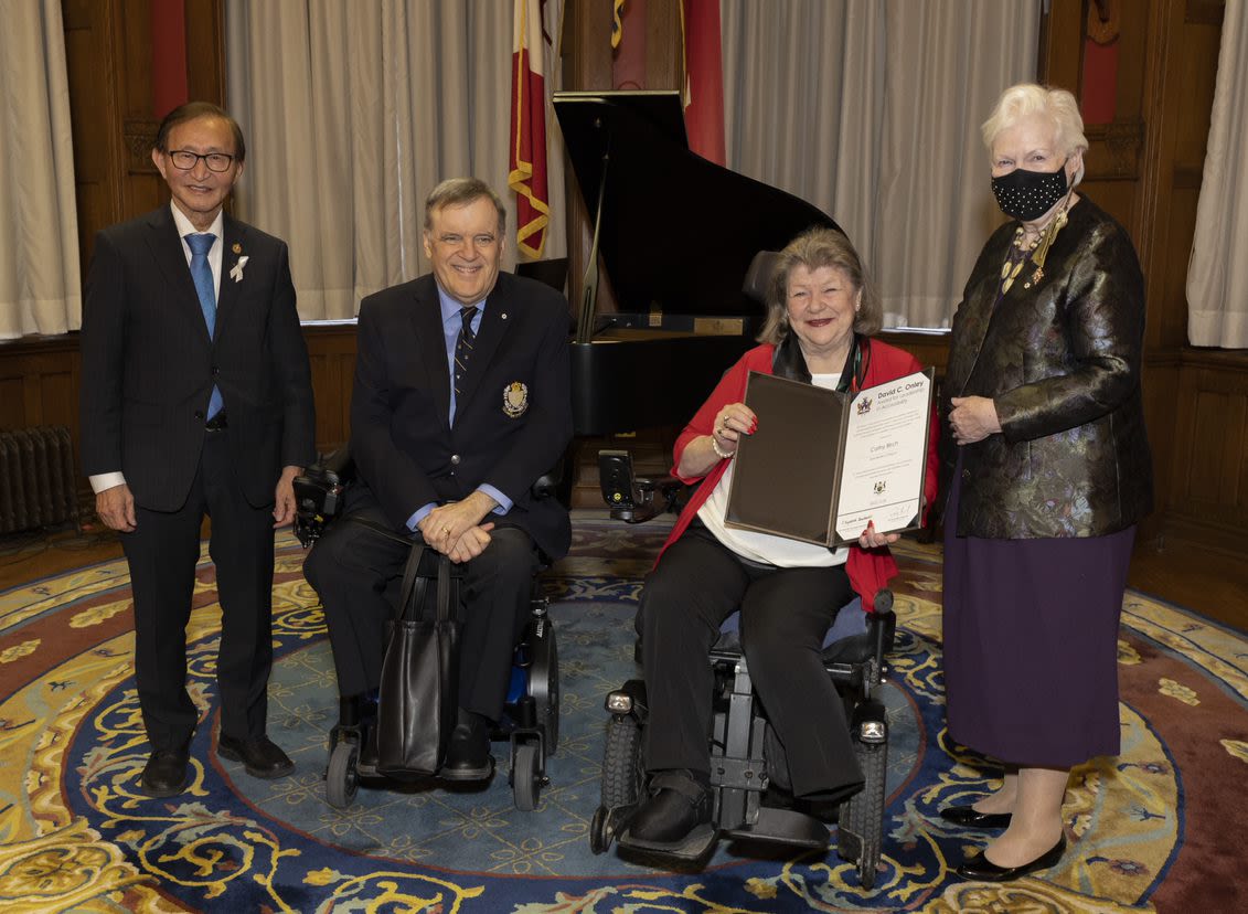 Photo of four people. The two people in the middle are seated in wheelchairs. One of the people in the middle is holding up a certificate. All people are smiling.