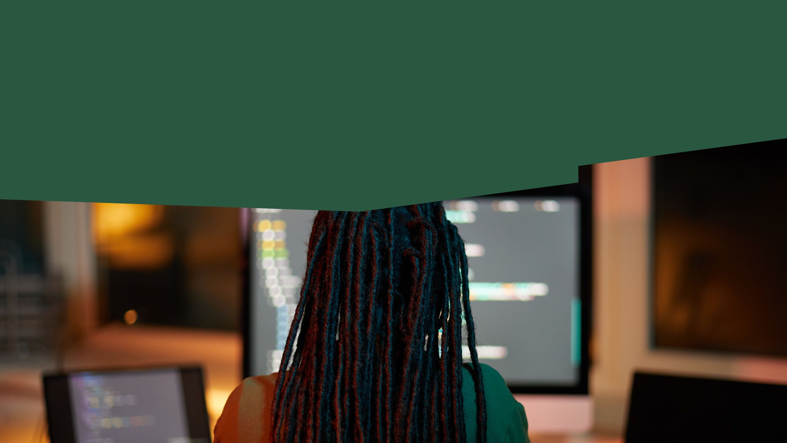 A person with braids sitting in front of a computer and tablet screen.