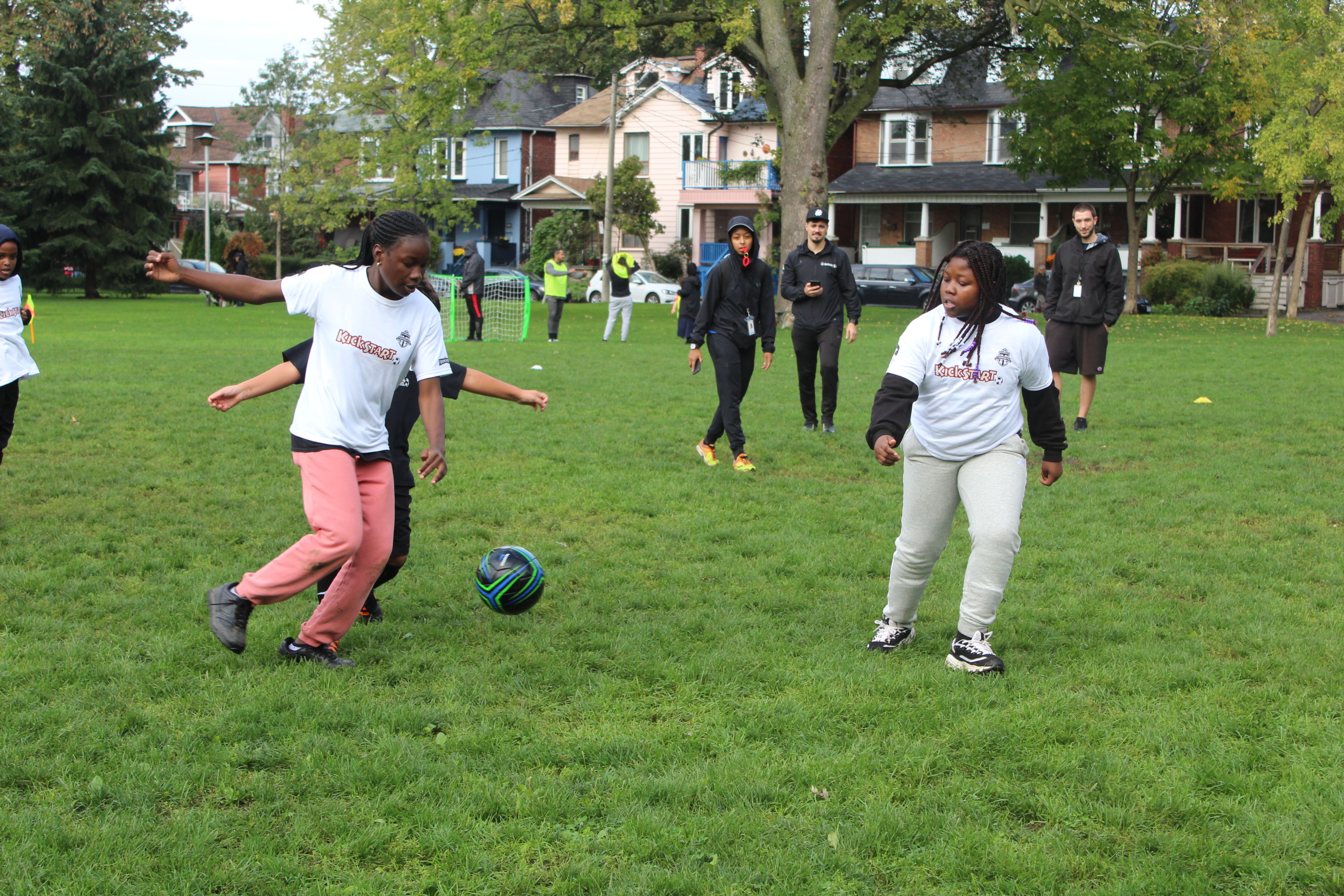 A group of children are playing soccer in a grassy field, coaches are standing in the distance watching them.