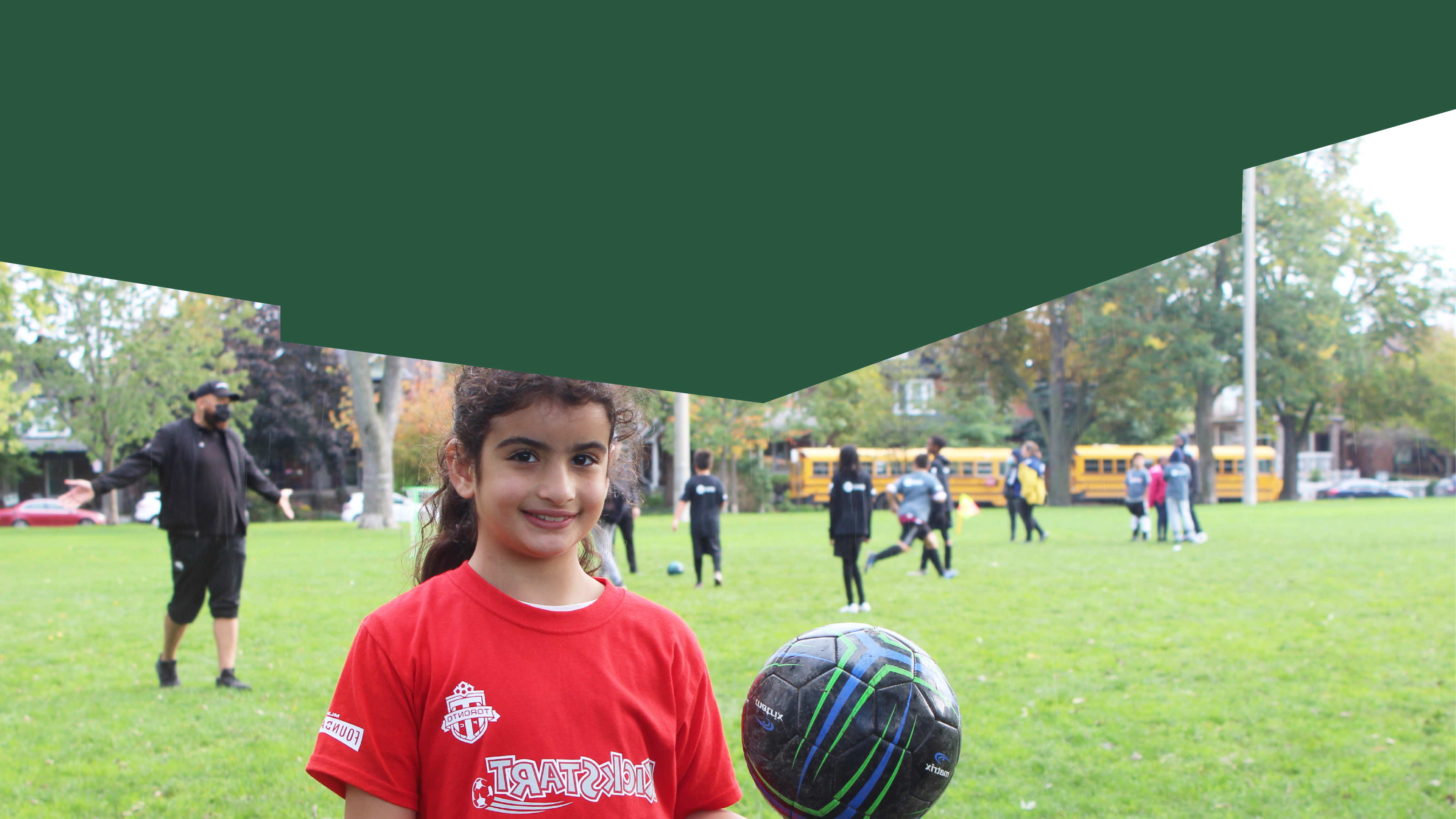 A child in a red shirt and pony tail is holding a soccer ball and smiling. In the background there are children playing soccer on a grassy field.