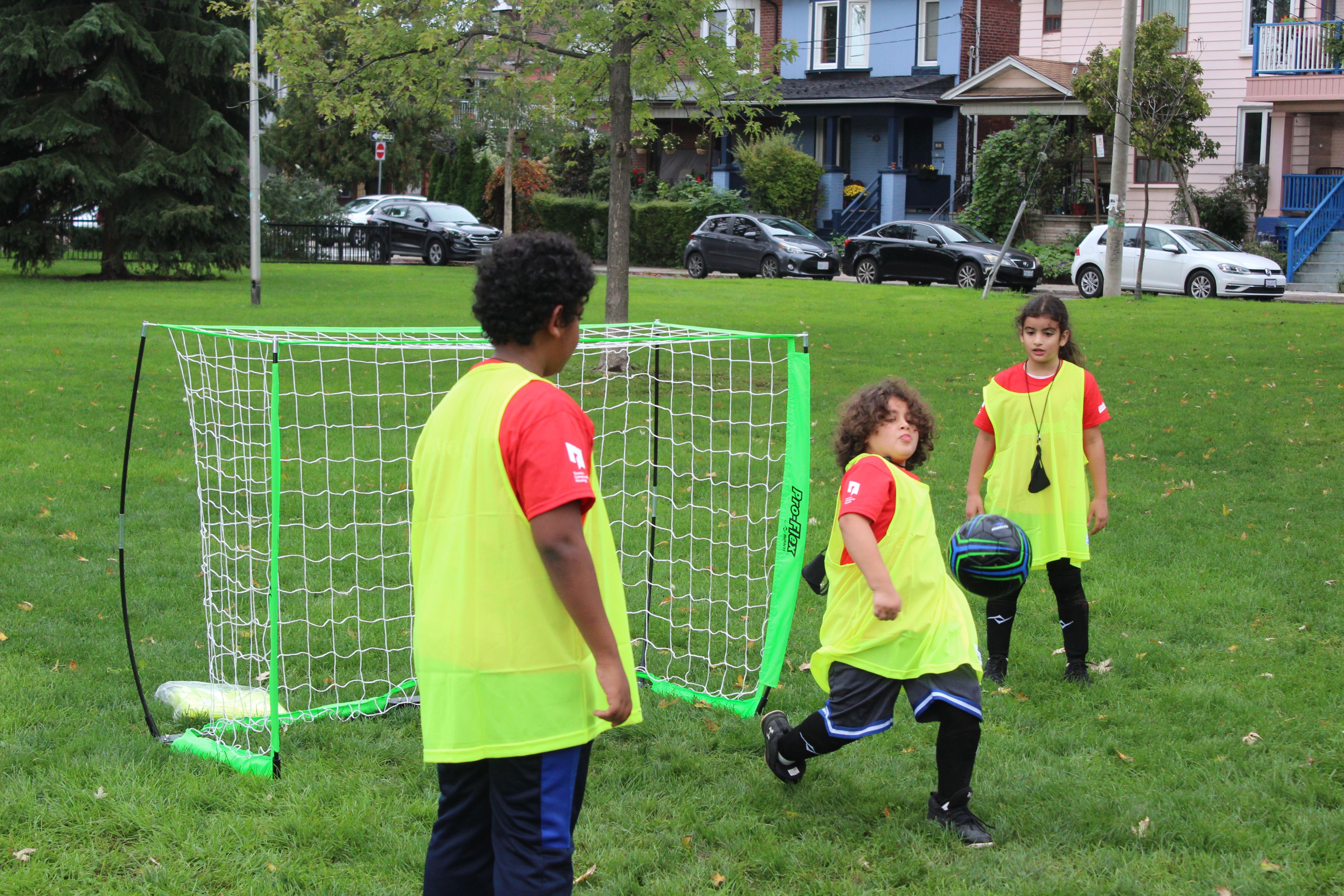 Three children in red t-shirts and fluorescent vests are playing soccer in a grassy field. 