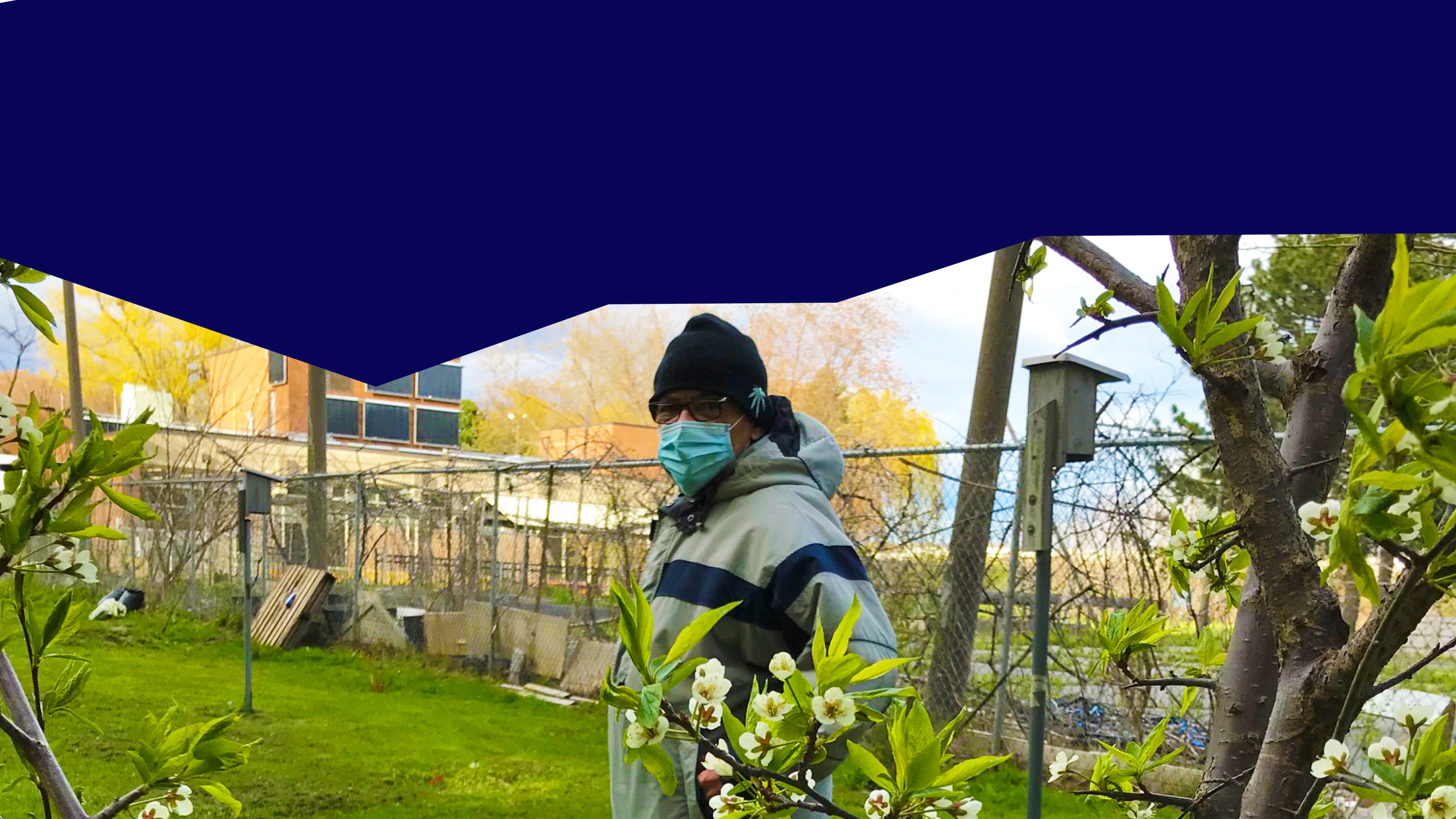 A person in glasses, a toque and a face mask is standing in a garden.