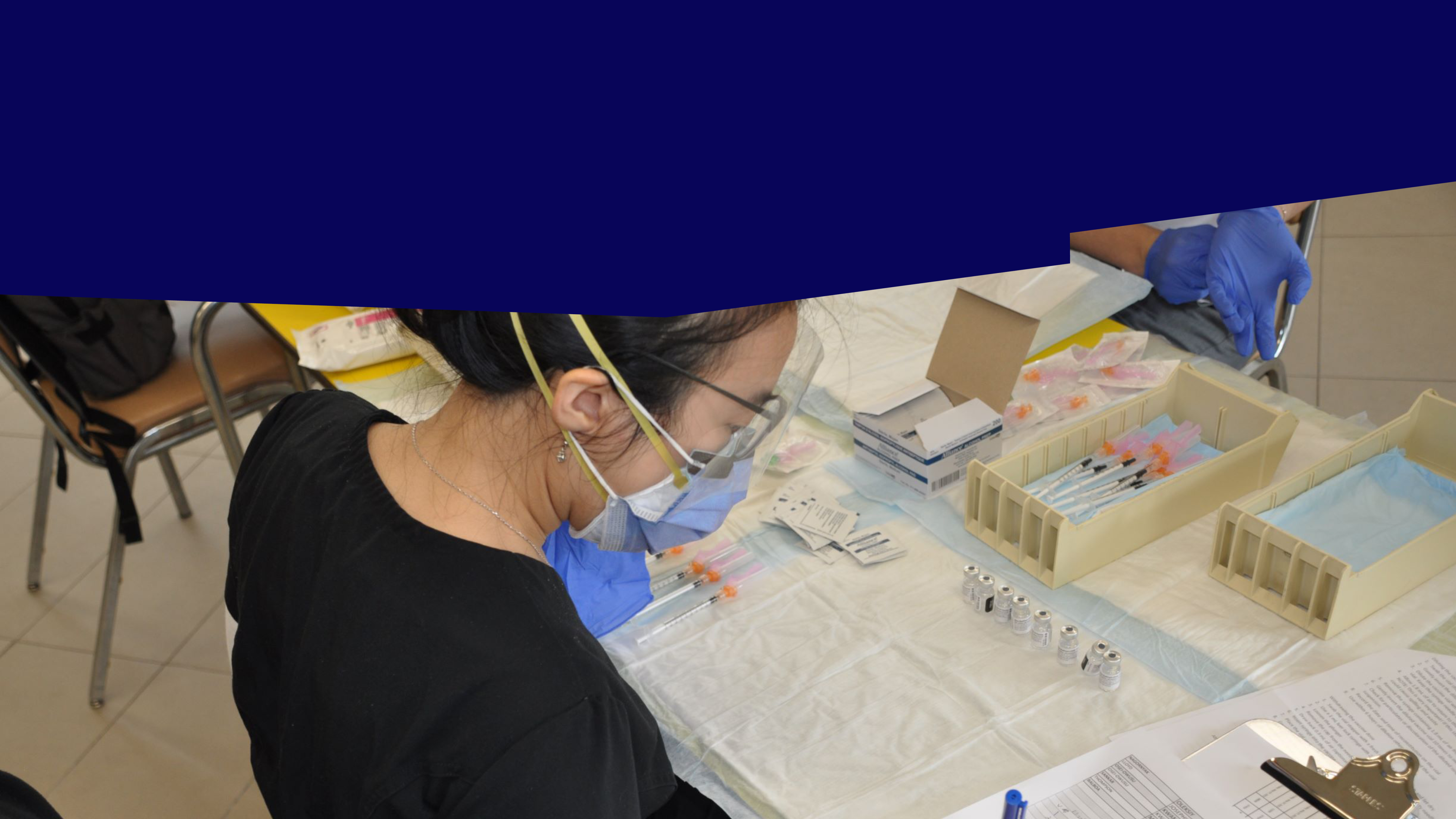 A healthcare worker sitting at a vaccination station with face masks on, the table in front of her has assorted medical supplies.