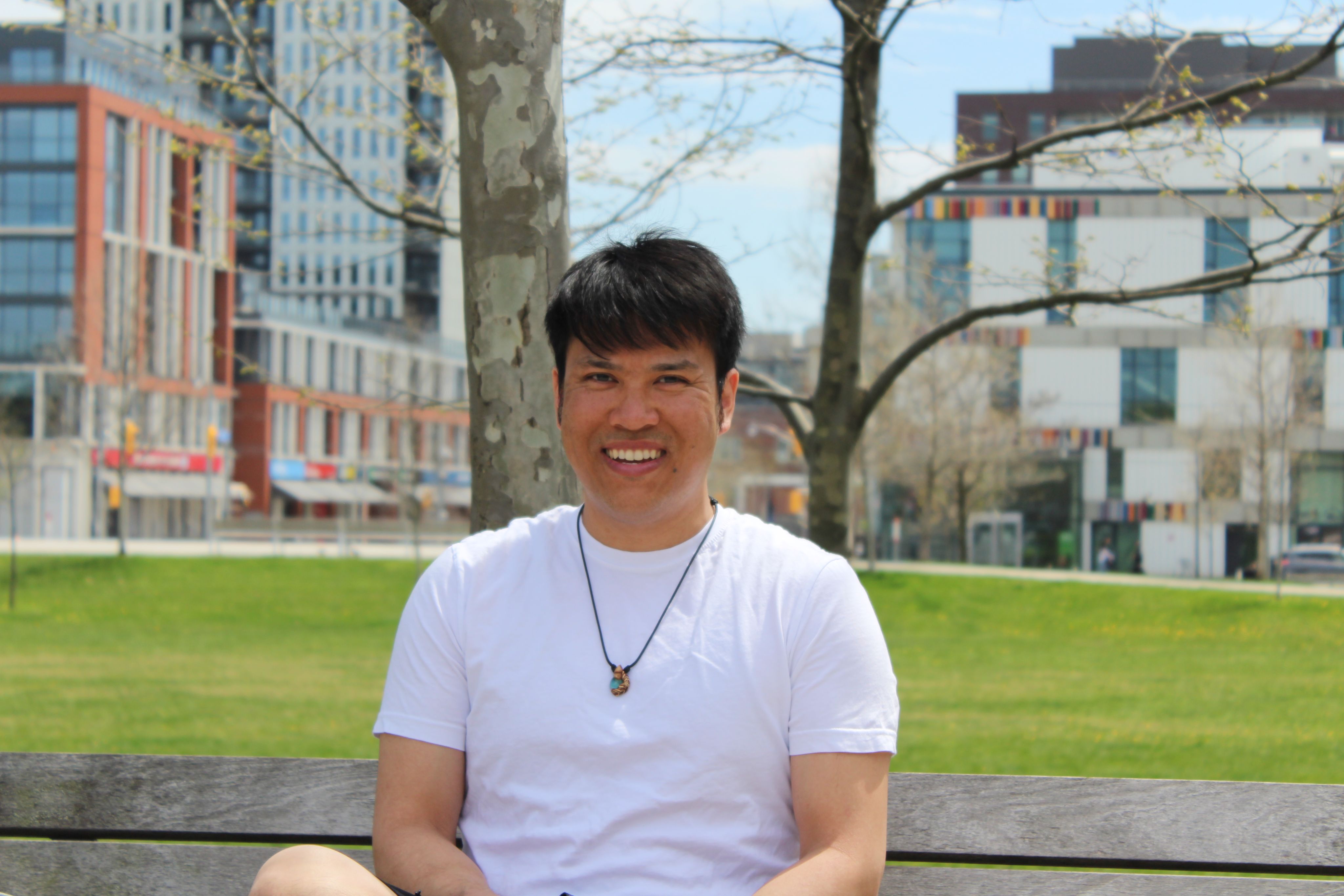 Tony Le sitting on a park bench with a tree and building in the background.
