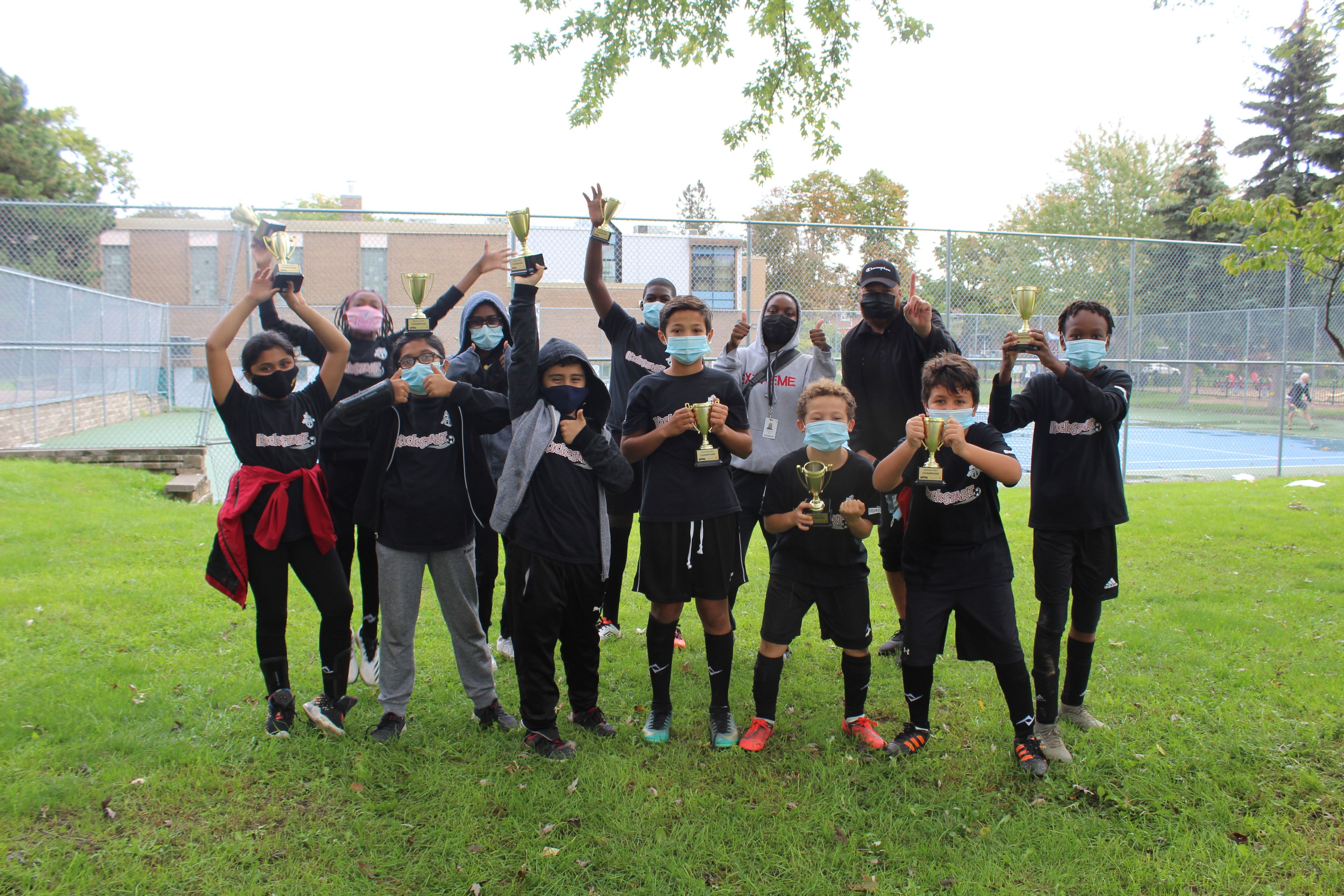 A group of children wearing face masks in grassy field celebrating and holding up trophies.