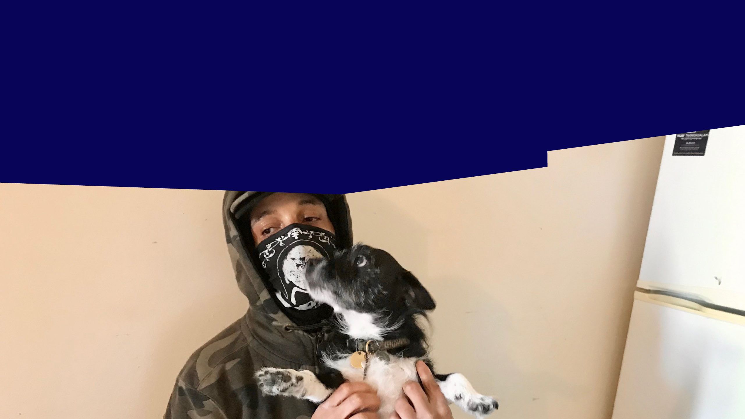 A person in a hooded sweatshirt and black facemask is holding up a black and white dog.