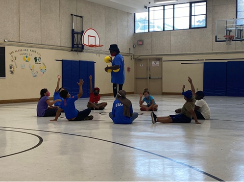 A group of youth sitting in a circle on an indoor basketball court have their hands raised, a coach stands in the middle holding basketballs.